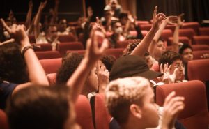 Fotografia de várias crianças sentadas no cinema com as mãos levantadas.