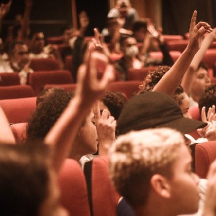 Fotografia de várias crianças sentadas no cinema com as mãos levantadas.