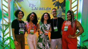 Fotografia de cinco pessoas, sendo quatro mulheres e um homem, posam sorridentes. Eles estão em pé, lado a lado, em frente a um banner com as palavras "FESTIVAL FALA! BELÉM DO PARÁ". O grupo está em um ambiente iluminado com luzes coloridas, todos usam crachás de identificação. No fundo, há plantas decorativas em ambos os lados.