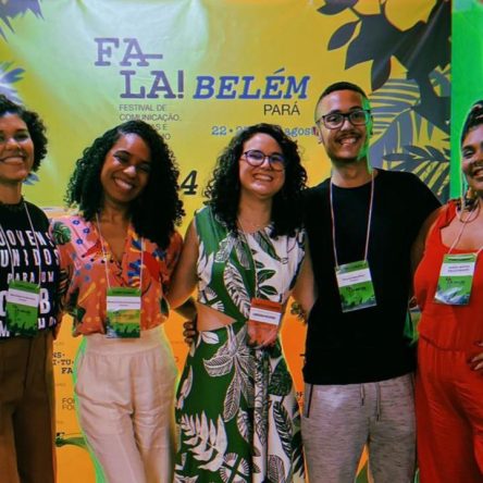 Fotografia de cinco pessoas, sendo quatro mulheres e um homem, posam sorridentes. Eles estão em pé, lado a lado, em frente a um banner com as palavras "FESTIVAL FALA! BELÉM DO PARÁ". O grupo está em um ambiente iluminado com luzes coloridas, todos usam crachás de identificação. No fundo, há plantas decorativas em ambos os lados.