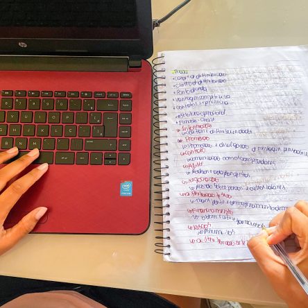 Fotografia de um laptop vermelho e preto com a mão esquerda em cima, enquanto escreve em um caderno espiral com a mão direita.