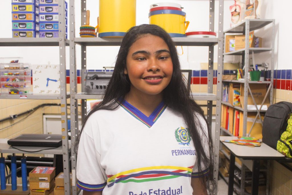 Fotografia uma menina negra, com cabelos longos e pretos, vestindo uma camisa branca com detalhes em azul, amarelo, verde e vermelho, que identifica a Rede Estadual de Ensino de Pernambuco. Ela está sorrindo e está em uma sala organizada, com estantes ao fundo contendo vários objetos, como caixas plásticas, instrumentos e materiais educativos.