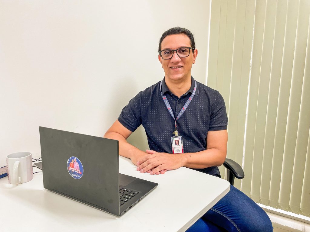 Fotografia de um homem branco, usando óculos e uma camisa polo preta com pequenos pontos brancos. Ele está sentado à mesa, em frente a um notebook. Ele sorri para a câmera e tem uma xícara ao lado, com persianas de fundo.