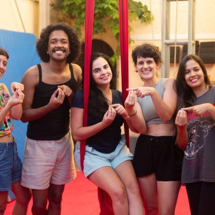 Um grupo de cinco pessoas está posando sorridentes em um ambiente interno com fundo de parede amarela e equipamentos de tecido acrobático e colchonetes azuis. Todas estão fazendo o sinal em libras Casulo.