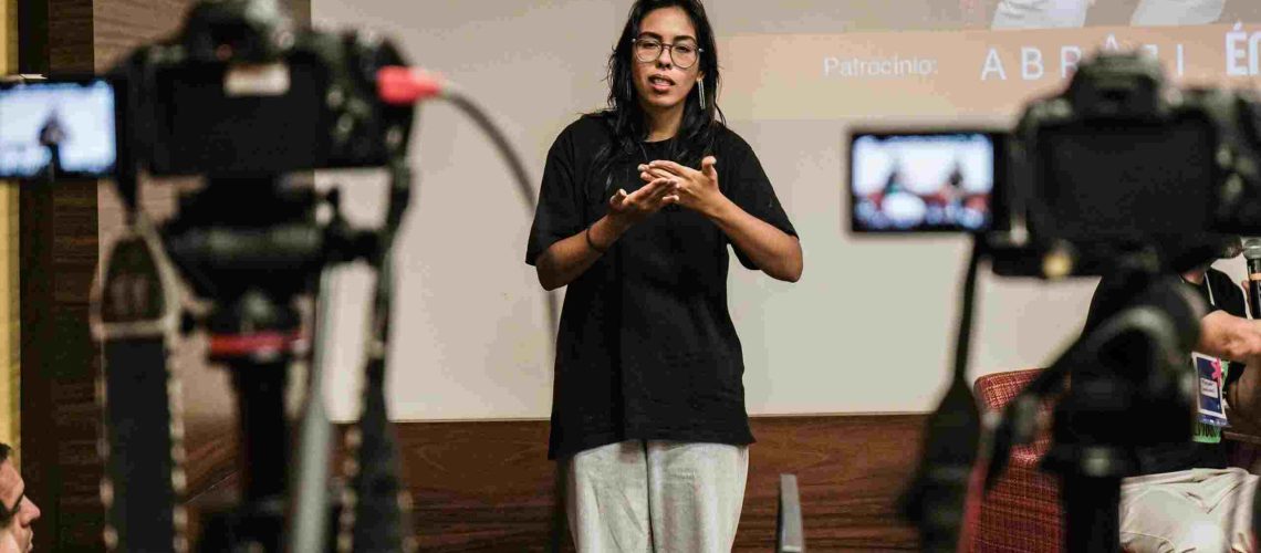 imagem mostra uma mulher de cabelo preto liso, usando óculos e blusa preta, sinalizando em Libras. Ao fundo, há um projetor e uma câmera desfocada.