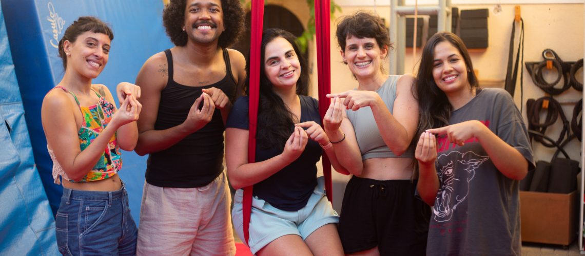 Um grupo de cinco pessoas está posando sorridentes em um ambiente interno com fundo de parede amarela e equipamentos de tecido acrobático e colchonetes azuis. Todas estão fazendo o sinal em libras Casulo.